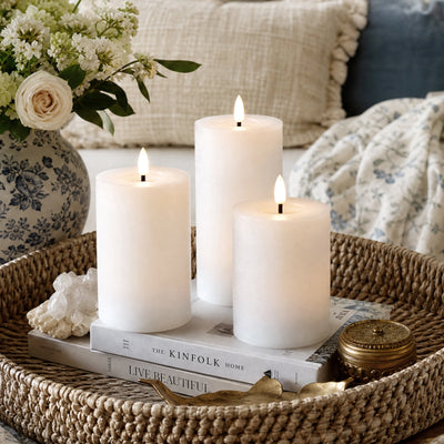 Three white candles on books with a vase of flowers and decorative items on a woven tray.