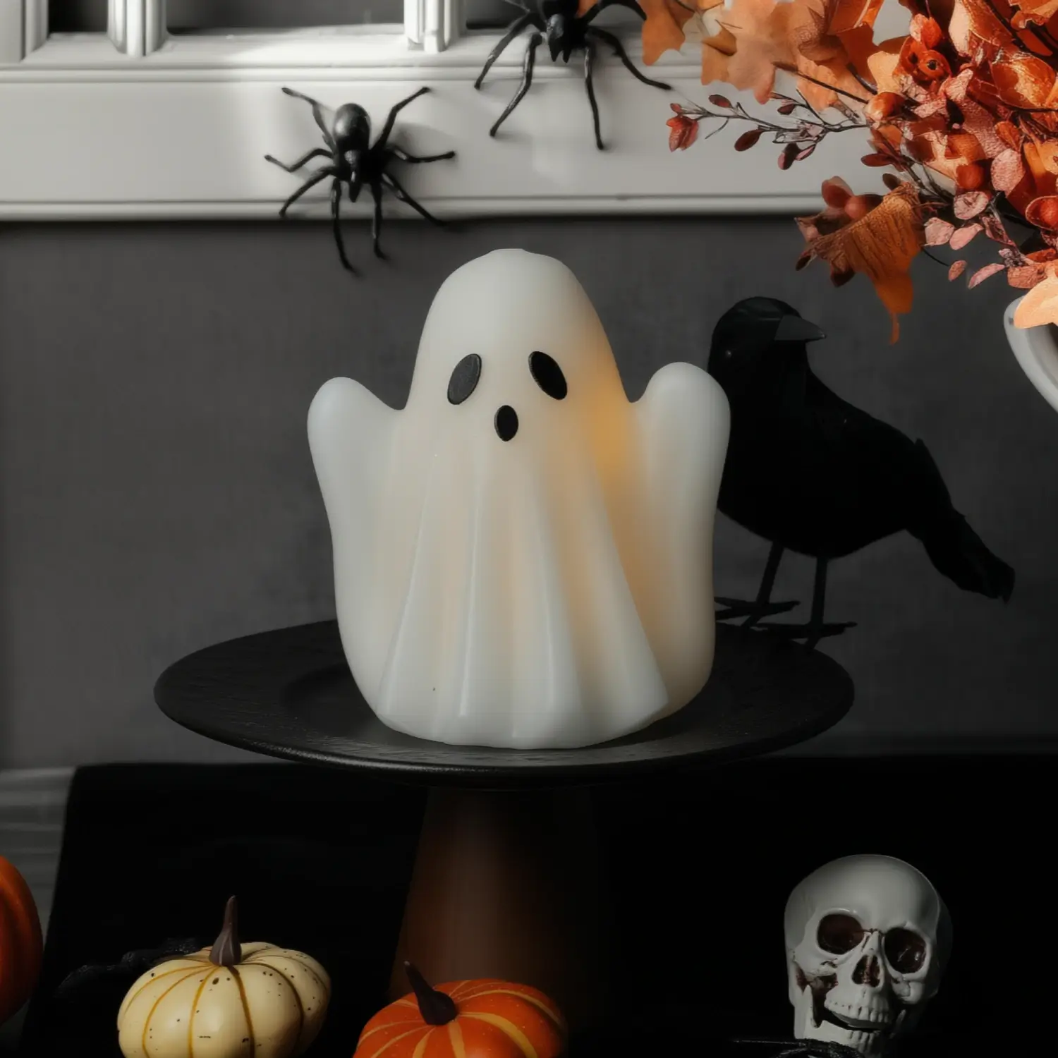 Ghost-shaped candle on a black stand with Halloween decorations including pumpkins, a skull, and spiders.