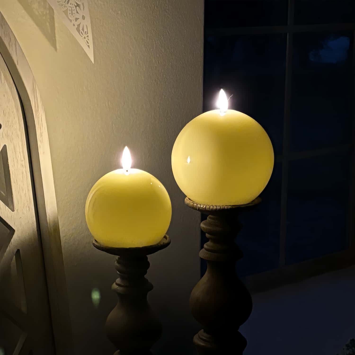 Two spherical yellow candles on dark stands against a neutral wall.