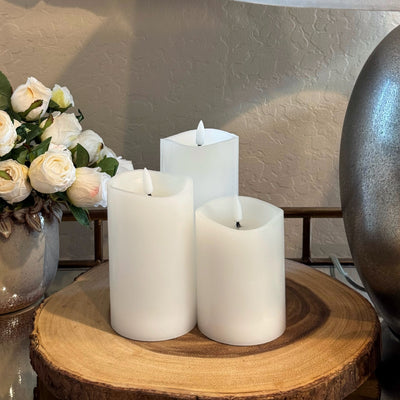 Three white candles on a wooden surface with a vase of flowers in the background.