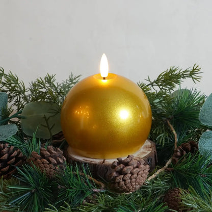 Gold spherical candle on a wooden base surrounded by greenery and pine cones.