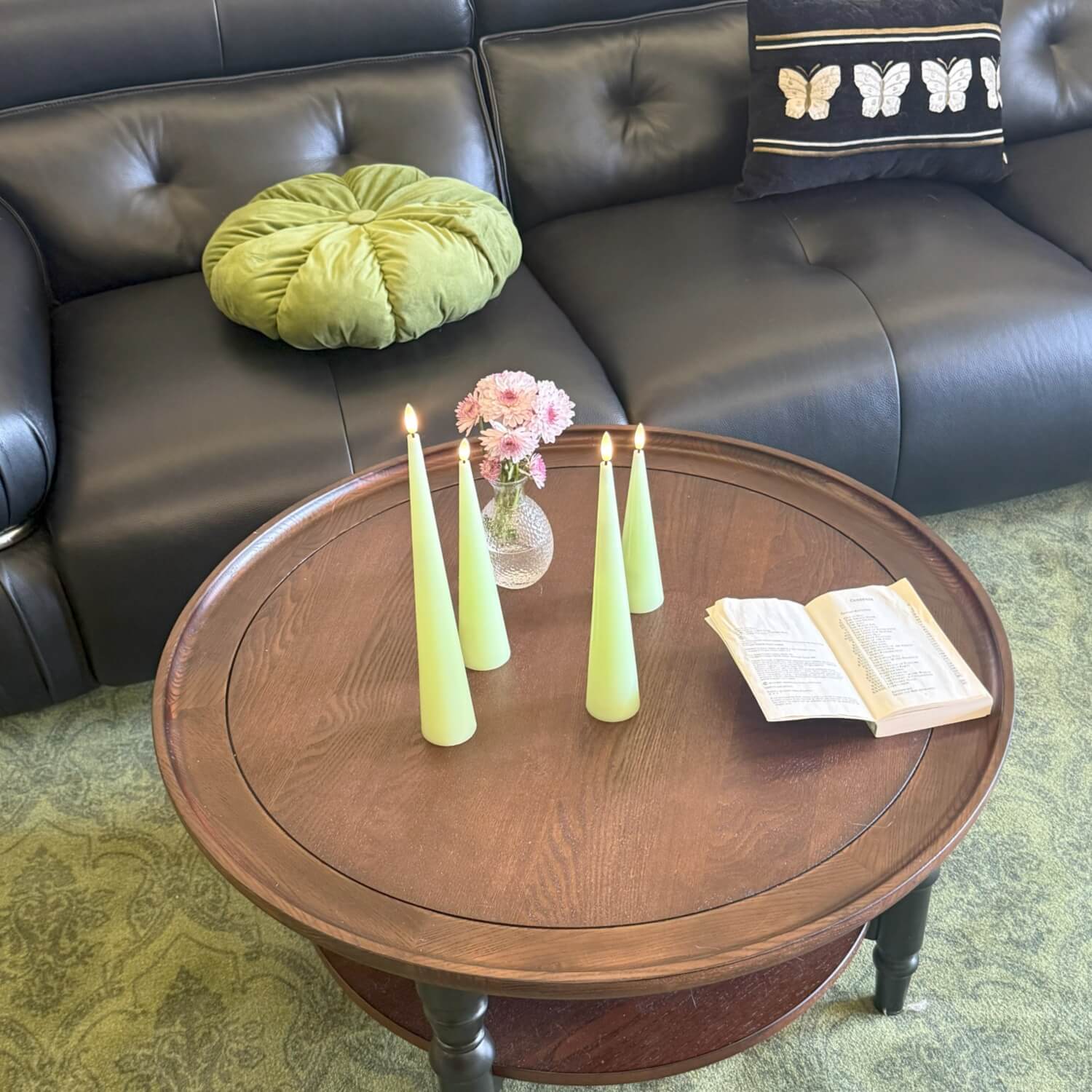 Round wooden coffee table with candles and a vase in front of a brown leather sofa.
