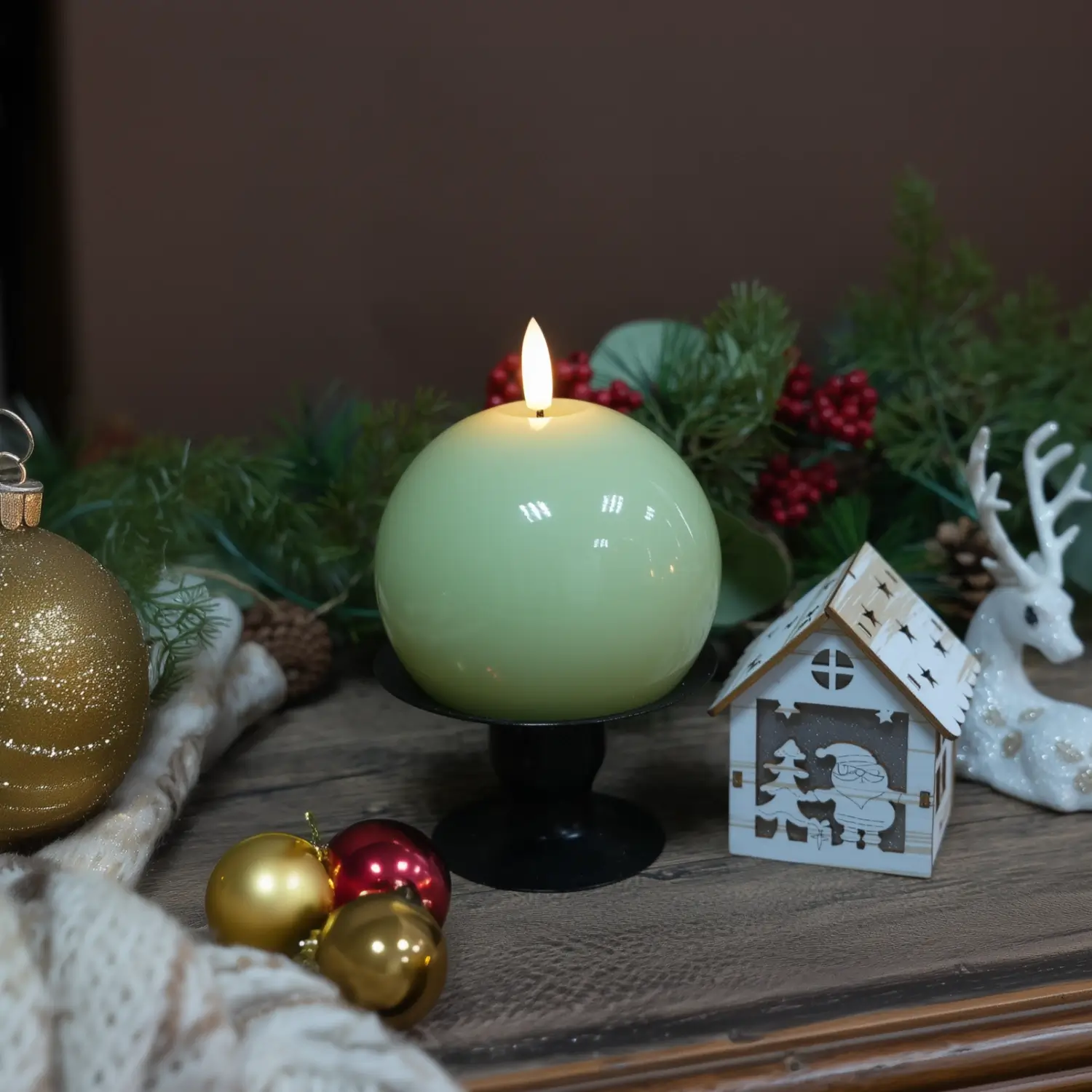 Green spherical candle with a wooden coaster on a table with Christmas decorations.
