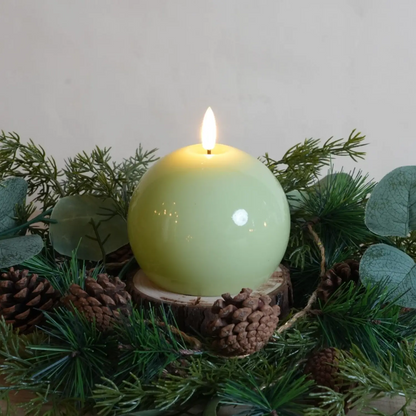 Green candle in a wooden holder surrounded by greenery and pinecones on a light gray background