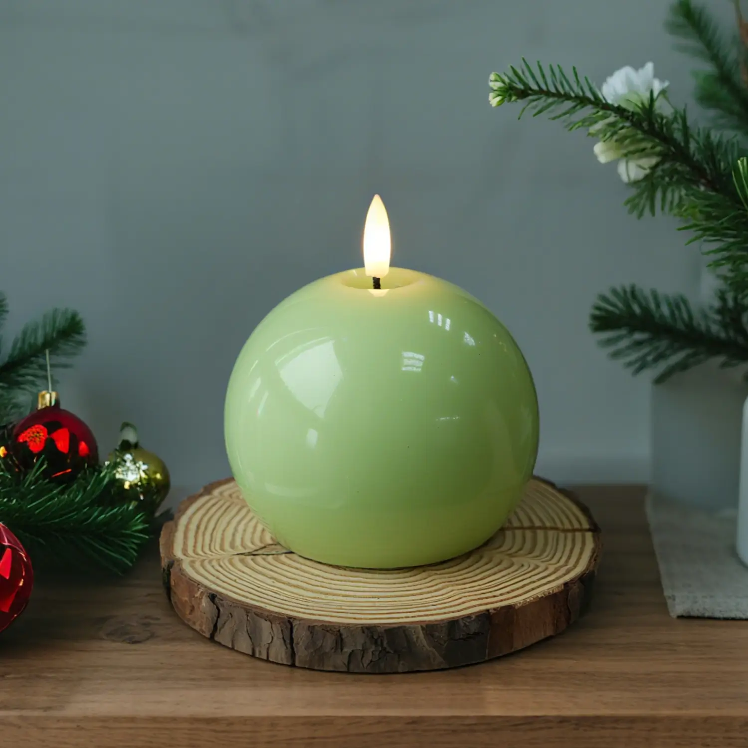 Green spherical candle with a wooden coaster on a table with Christmas decorations.