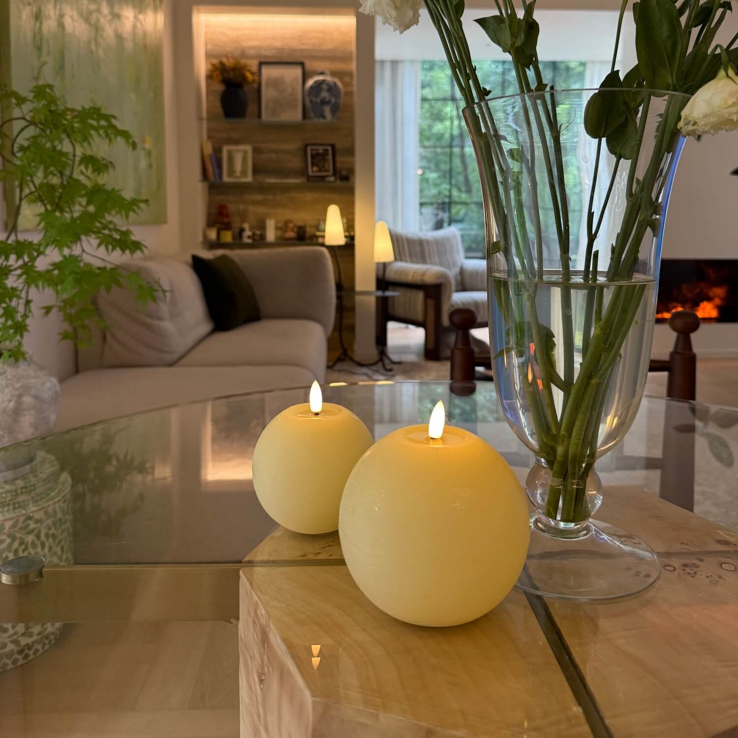Two lit candles on a glass coffee table with a vase of flowers in the background.