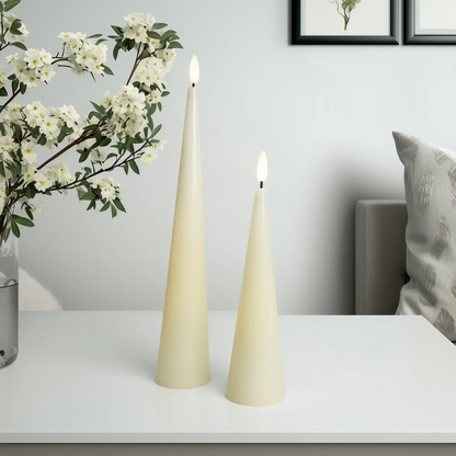 Two cone-shaped candles on a white surface with a vase of flowers and a pillow in the background.