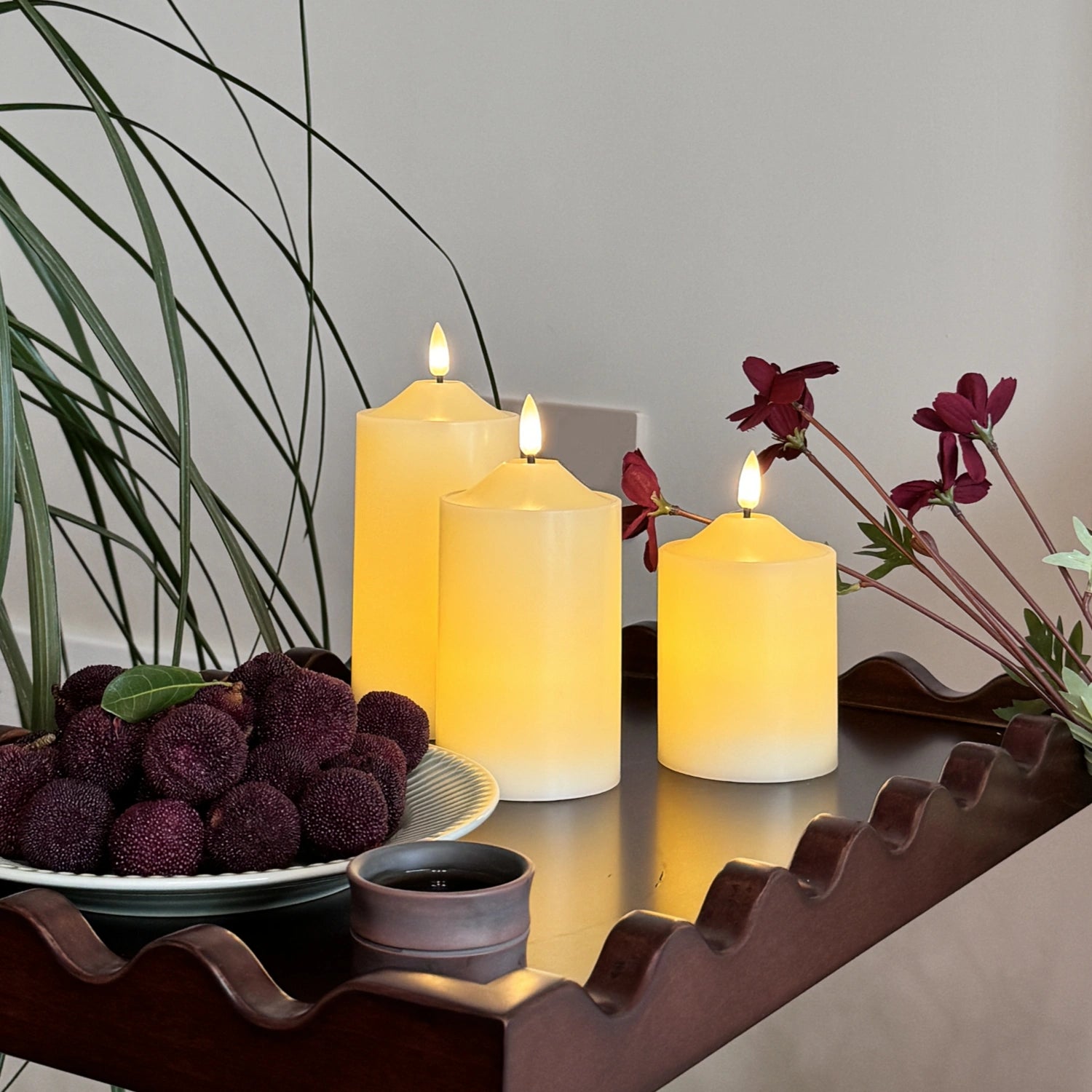 Three lit candles on a decorative stand with purple fruits and flowers.