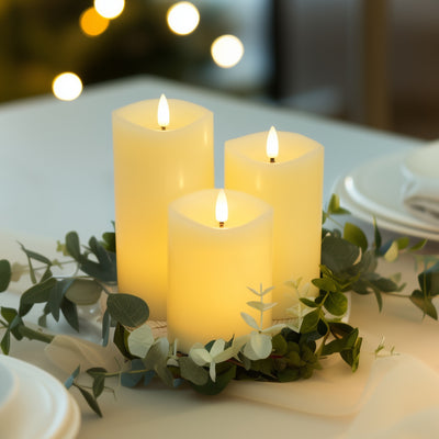 Three lit candles on a table with greenery and blurred lights in the background