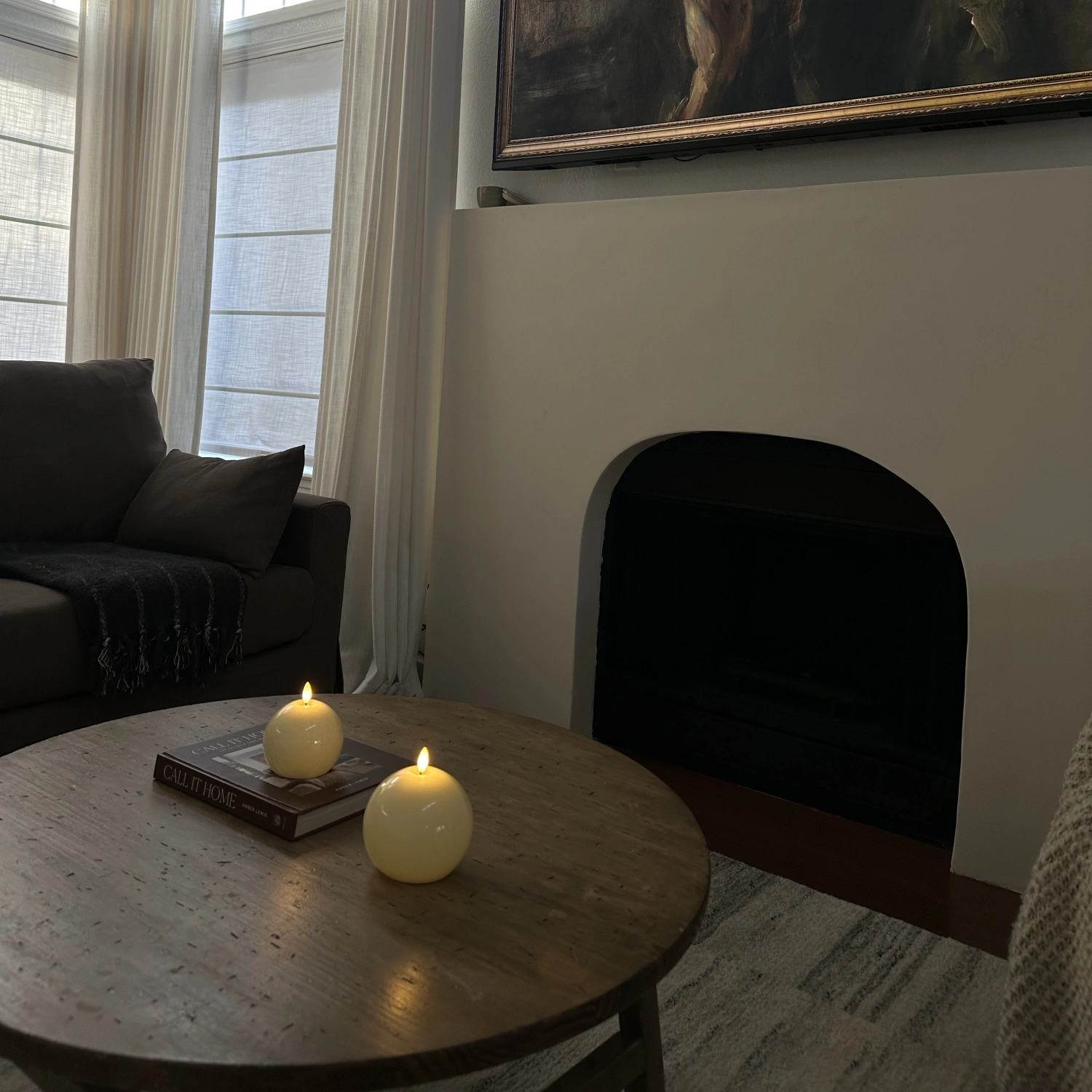 Living room with a round wooden table featuring two lit candles and a book, next to a dark sofa and a fireplace.