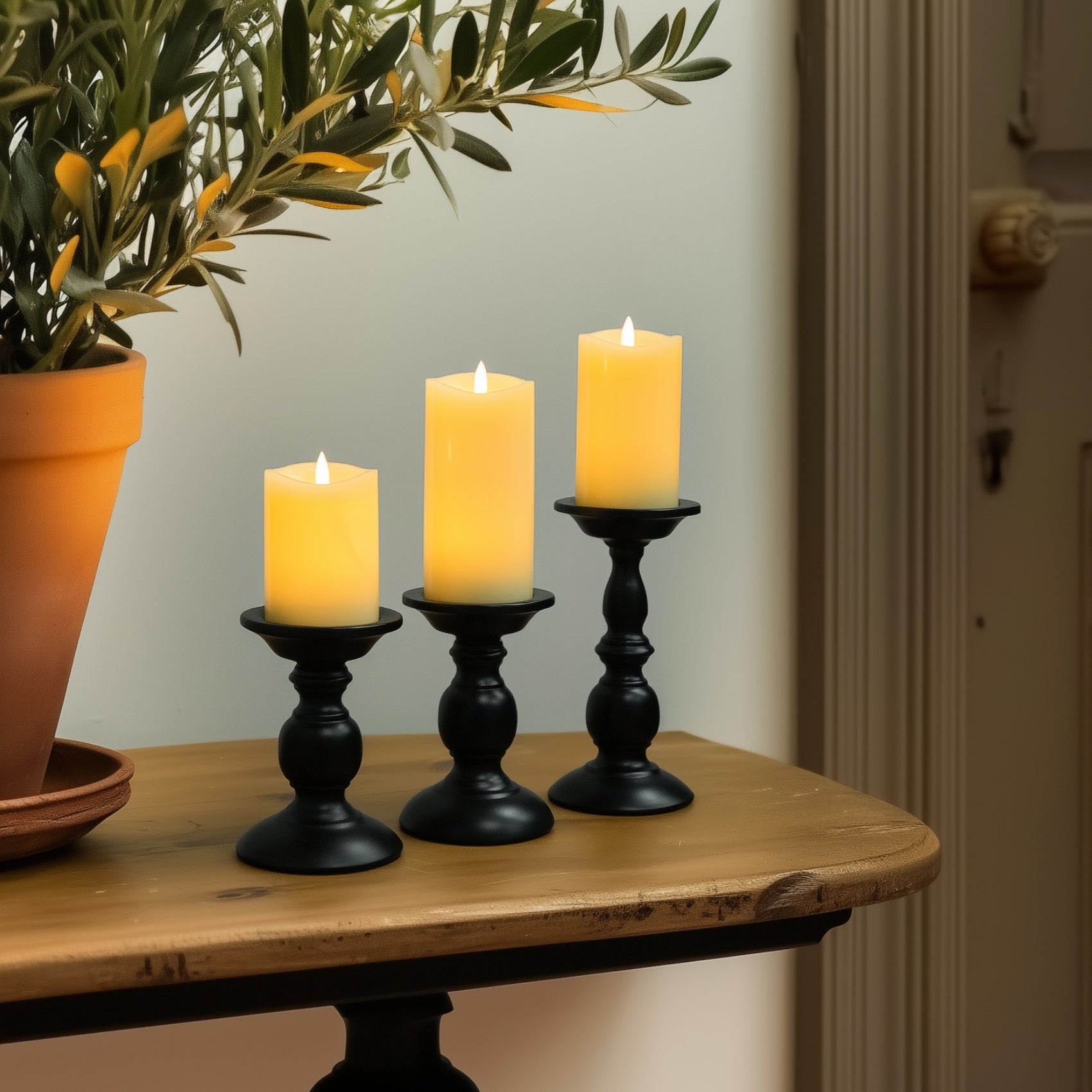 Three lit candles in black candlesticks on a wooden surface with a plant in the background.