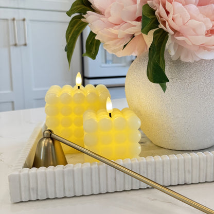Two lit candles in a decorative holder with a vase of pink flowers in the background.