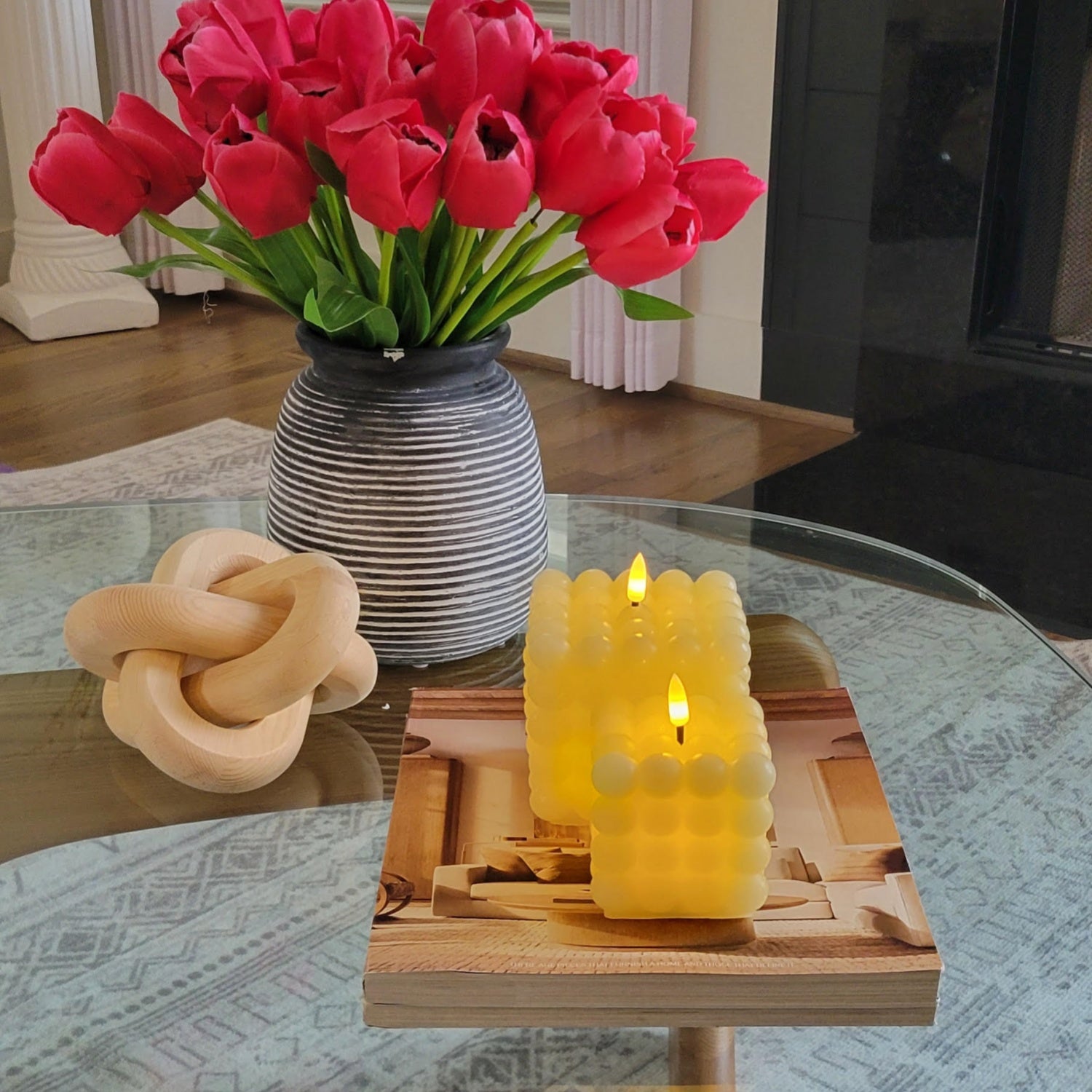 Two lit candles on a wooden coaster with a vase of red tulips in the background.