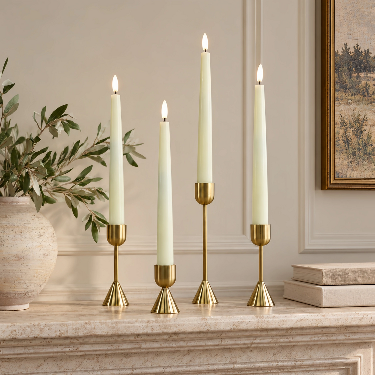 Four cream taper candles in gold candlesticks arranged on a stone mantel in a styled interior setting with a vase of greenery and framed wall art.