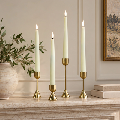 Four cream taper candles in gold candlesticks arranged on a stone mantel in a styled interior setting with a vase of greenery and framed wall art.