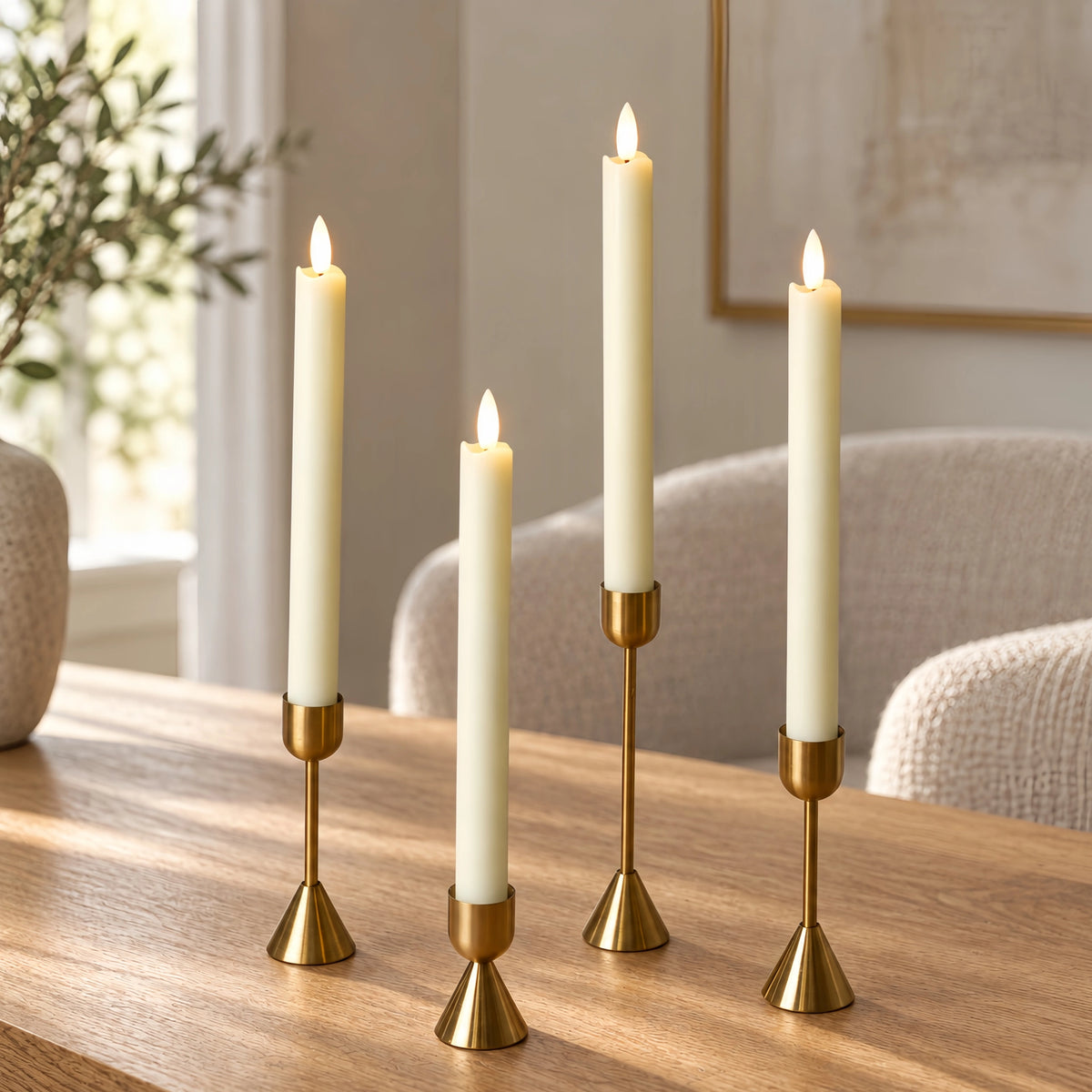 Four cream pillar candles in varying heights, each lit and displayed in a gold metal holder on a wooden table in a softly blurred living room setting.