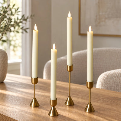 Four cream pillar candles in varying heights, each lit and displayed in a gold metal holder on a wooden table in a softly blurred living room setting.