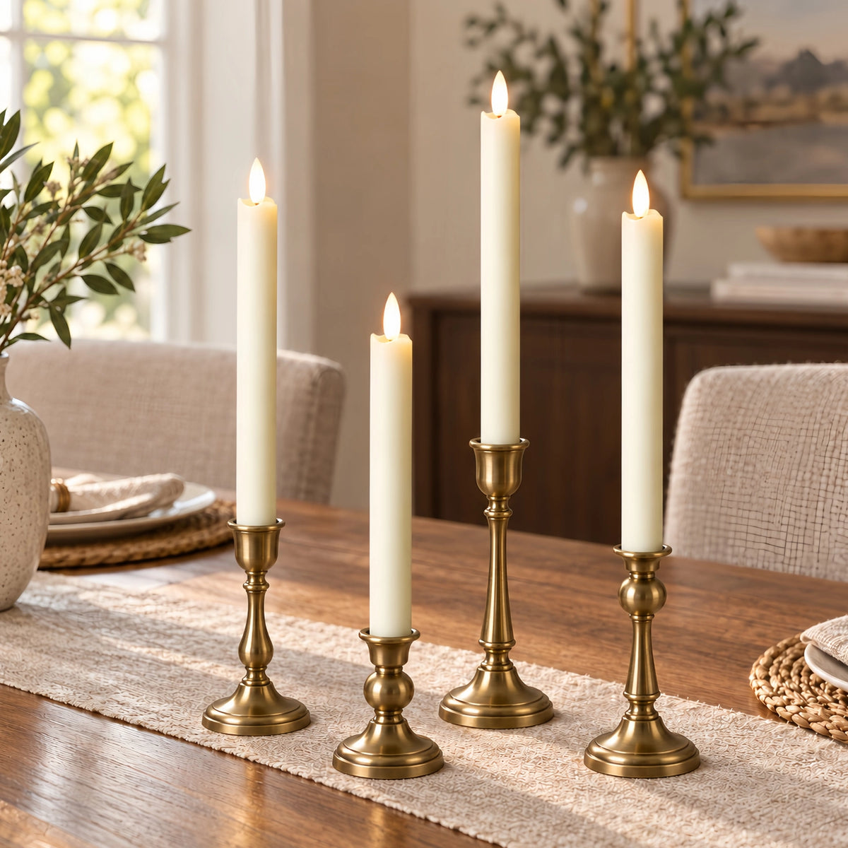 Four cream pillar candles in matching brass candlesticks arranged on a woven table runner in a warm, softly lit dining room.
