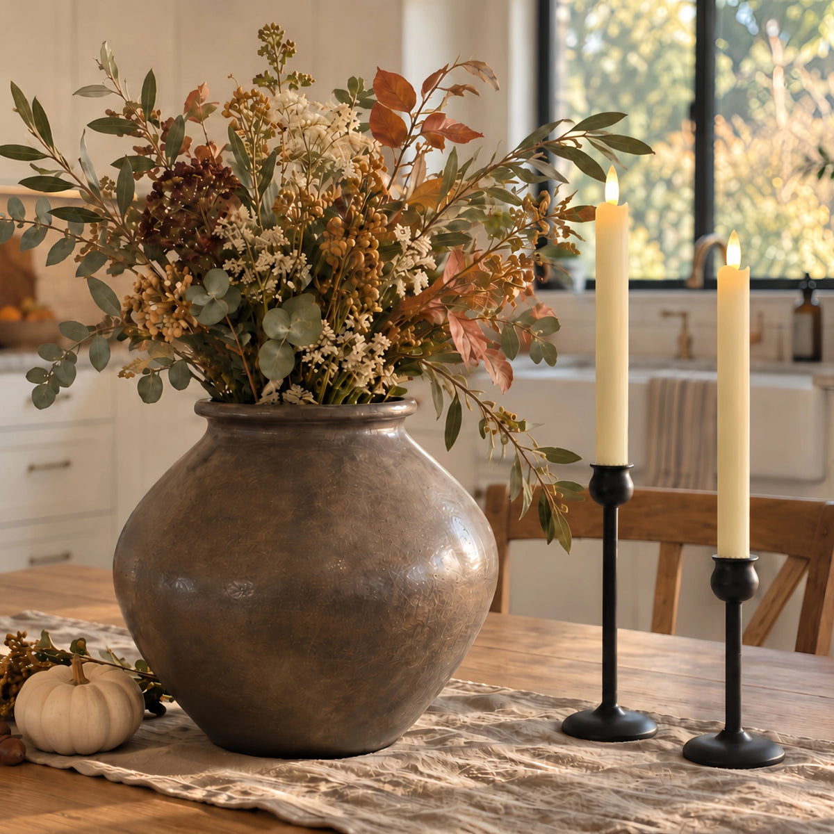 A large round metallic bronze-brown vase filled with a dried floral arrangement in muted beige, brown, and green tones, set on a dining table beside two lit white taper candles in black holders, with a softly blurred kitchen background.