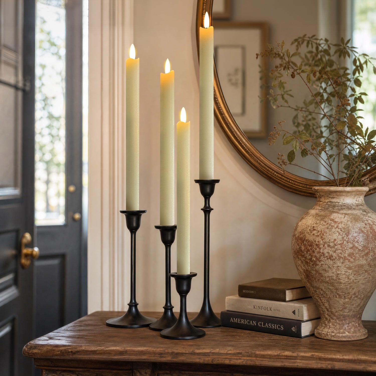 Four black metal taper candlesticks holding lit cream pillar candles, styled on a wooden console table beside a large textured vase, books, and a mirror in an indoor room.