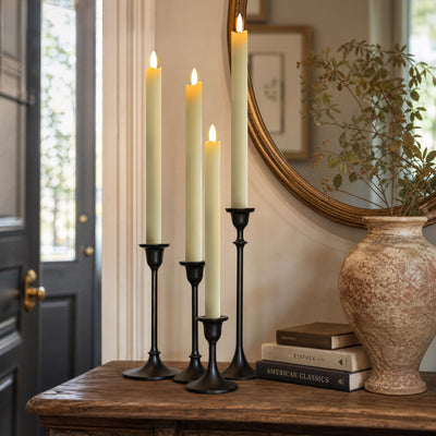 Four black metal taper candlesticks holding lit cream pillar candles, styled on a wooden console table beside a large textured vase, books, and a mirror in an indoor room.