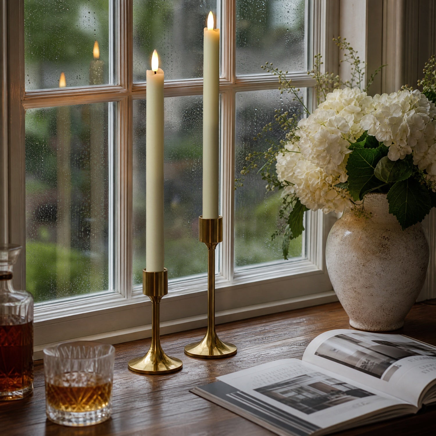 Two lit cream pillar candles in gold-toned holders on a wooden windowsill, with a vase of white flowers, an open book, and a glass of amber liquid nearby.