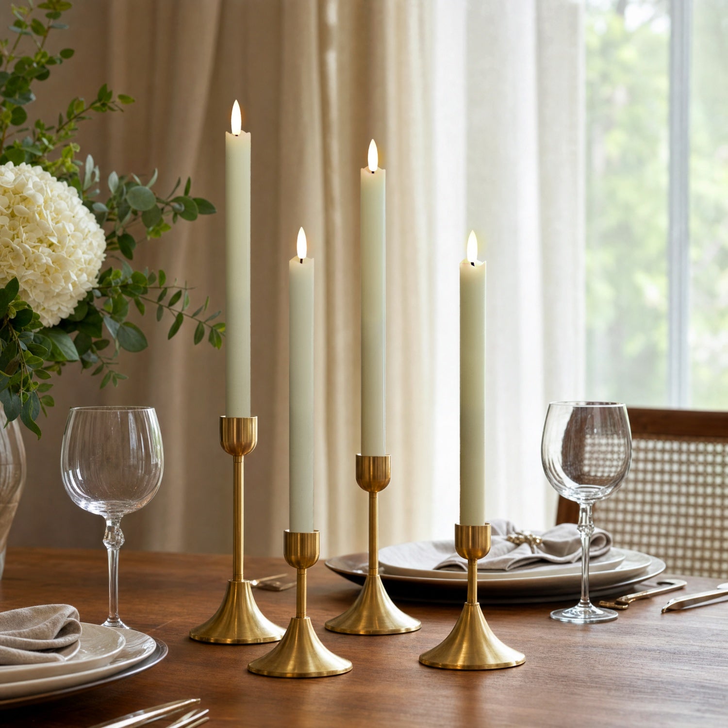 Four white taper candles in gold candlesticks arranged on a wooden dining table, with plates, wine glasses, and a floral centerpiece in a softly lit room.
