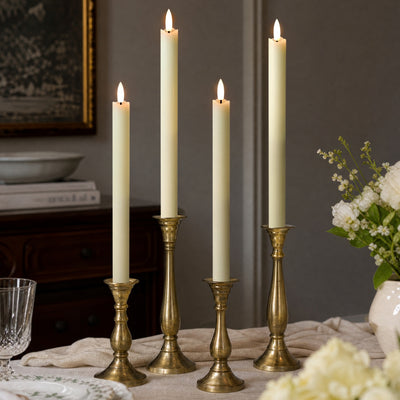 Three lit cream taper candles in ornate gold candlesticks arranged on a table in a styled interior setting.