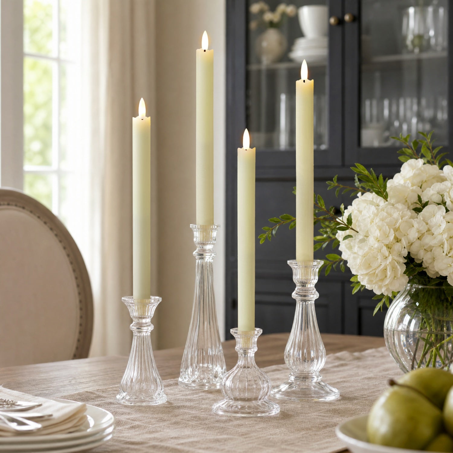Four cream pillar candles in clear glass candlesticks arranged on a dining table, with flowers and green pears in a softly lit room.