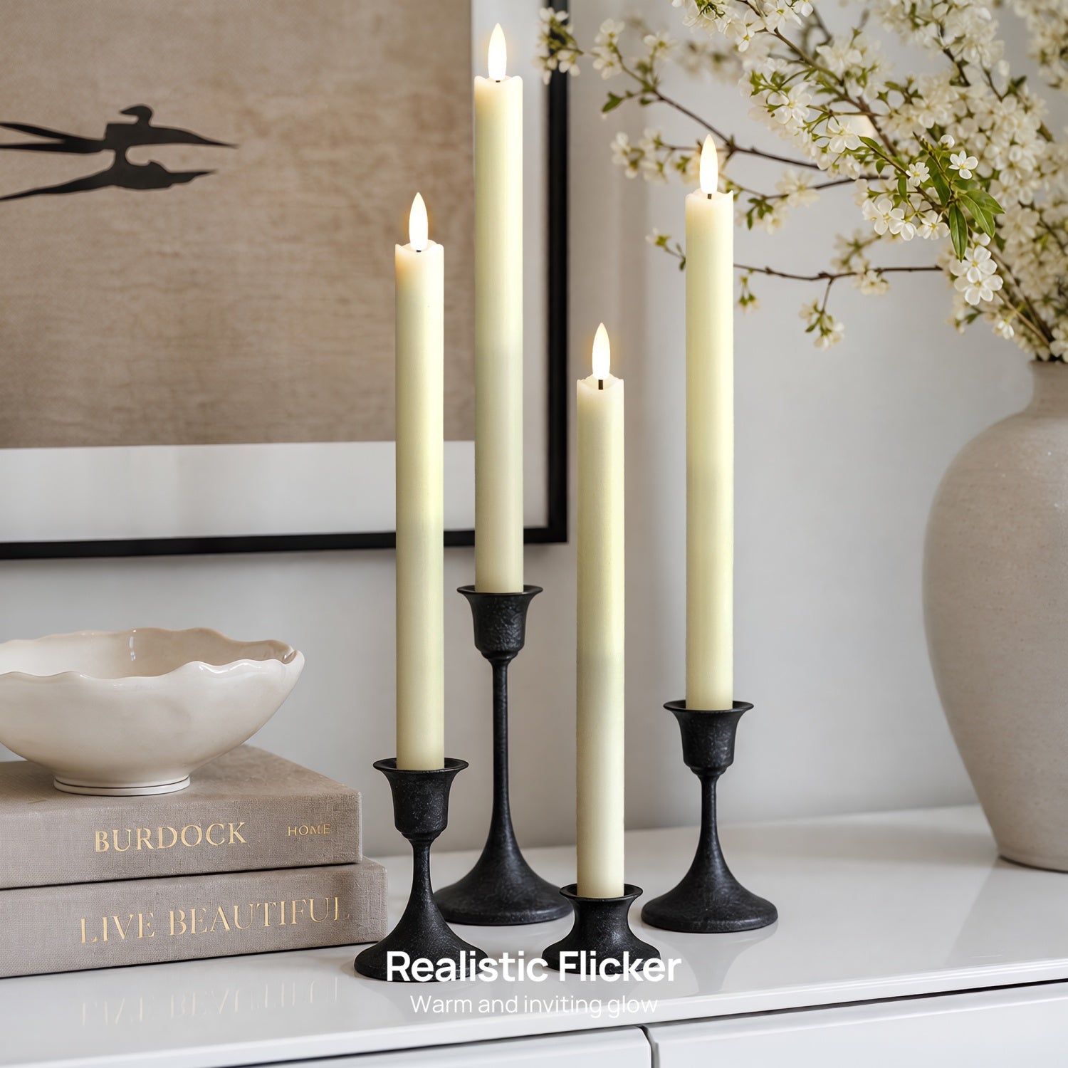 Three black candlestick holders with cream pillar candles lit, styled on a white surface with books, a bowl, framed wall art, and a vase of white blossoms in a bright interior setting.#color_rustic-ivory
