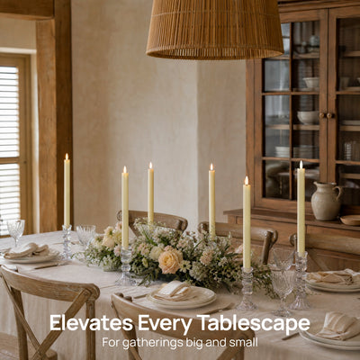 A styled dining table in a rustic indoor room with a floral centerpiece, multiple lit taper candles in glass holders, white plates, folded napkins, and wooden chairs. A large woven pendant light hangs above, and a wooden cabinet is visible in the background.