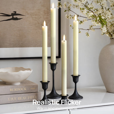 Three black metal candlesticks holding cream pillar candles with realistic flickering flames, styled on a white surface with books, a bowl, framed wall art, and a vase of white blossoms in a bright interior setting.