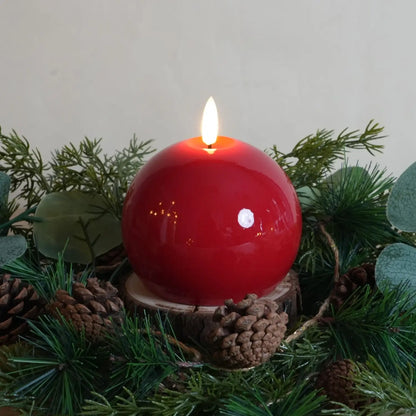 Red candle in a decorative holder surrounded by greenery and pinecones on a light background