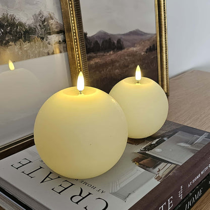 Two spherical candles with flame on a bookshelf with a framed landscape painting in the background.