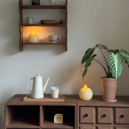 Wooden shelf with a teapot, cups, and candles, next to a potted plant on a wooden cabinet.
