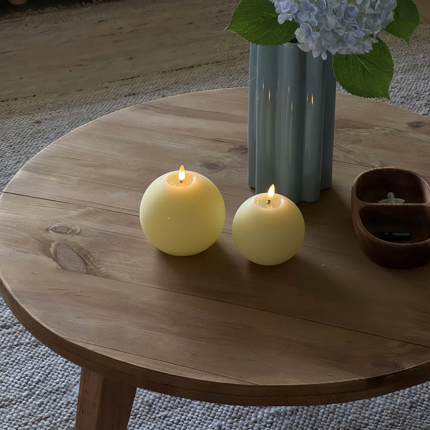 Two lit candles on a wooden table with a vase of flowers in the background.