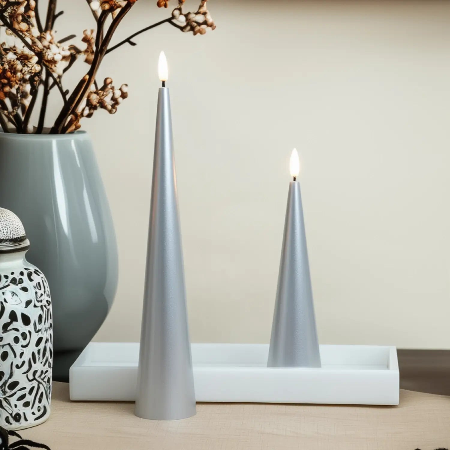 Two lit silver candles on a white surface with a vase and decorative items in the background.