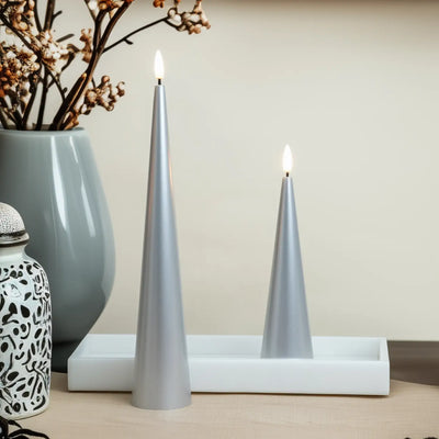 Two lit silver candles on a white surface with a vase and decorative items in the background.