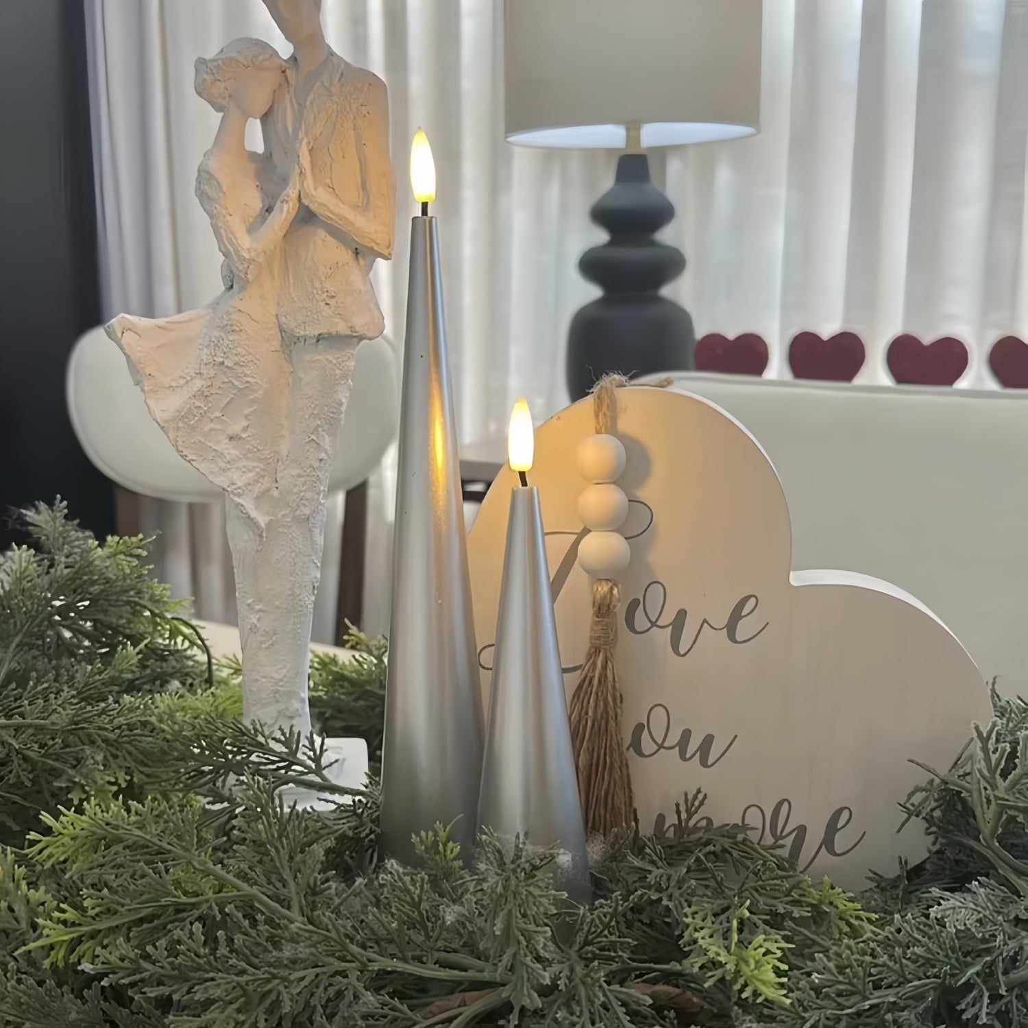 Two lit silver candles on a white surface with a vase and decorative items in the background.