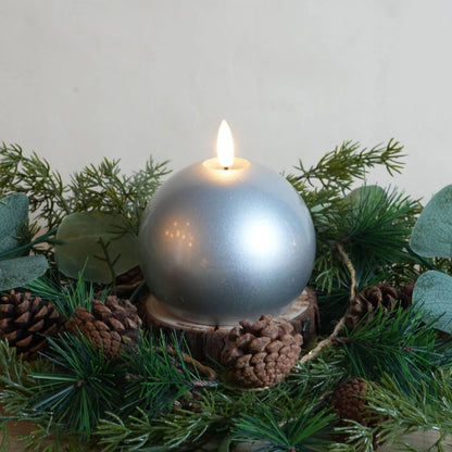 Silver ball candle surrounded by greenery and pine cones on a neutral background