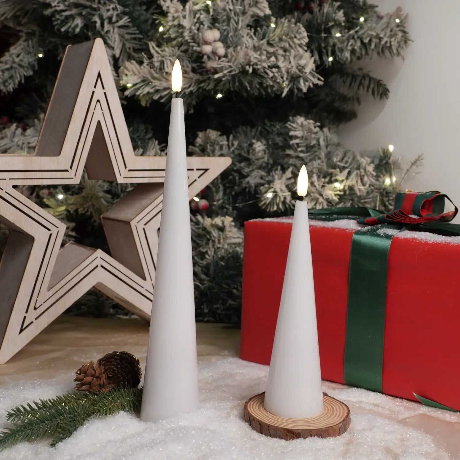 Two white candles on a wooden stand with a Christmas tree and gift in the background.