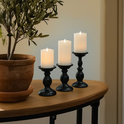 Three lit candles in black candlesticks on a wooden table with a potted plant.