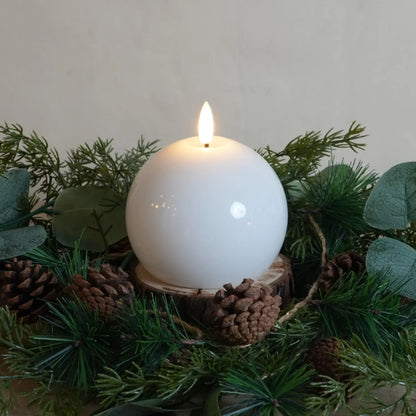 White candle on a wooden base surrounded by greenery and pine cones.