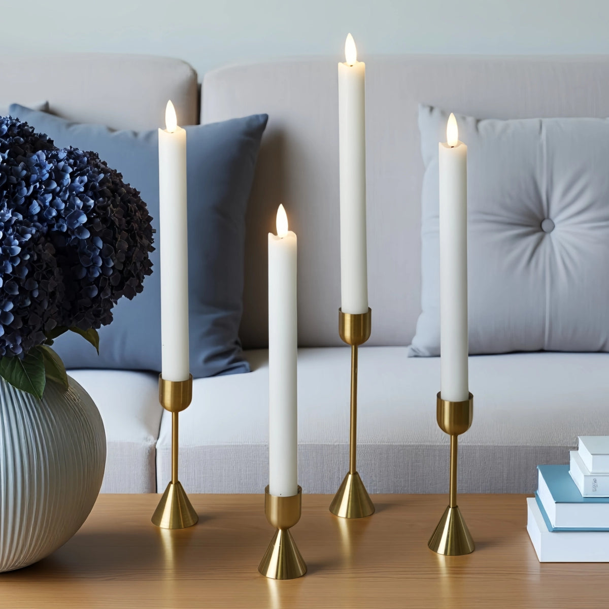 Four white taper candles in gold metal holders arranged on a wooden table in front of a light gray sofa with blue pillows. A white vase with dark blue hydrangeas sits on the left, and a stack of books is on the right.