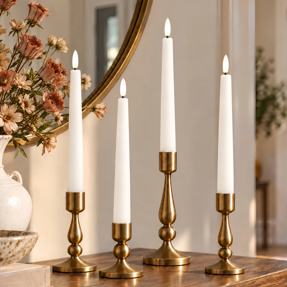 Four white taper candles in ornate gold candlesticks arranged on a wooden table in a warmly lit interior setting with a mirror and flowers in the background.