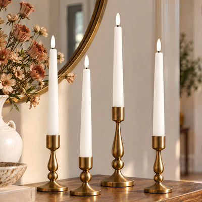 Four white taper candles in ornate gold candlesticks arranged on a wooden table in a warmly lit interior setting with a mirror and flowers in the background.