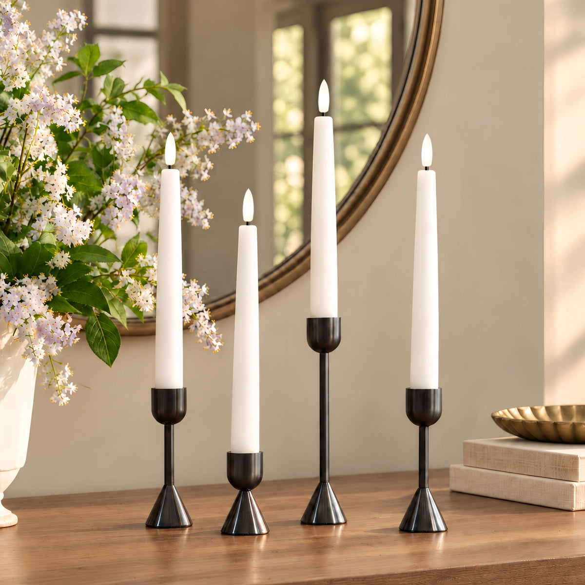 Four white taper candles in black metal holders arranged on a wooden table in a styled interior setting with a mirror, flowers, and books.