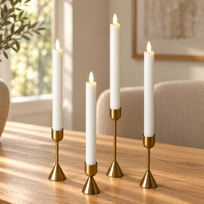 Four white taper candles in slim gold metal holders arranged on a wooden table in a softly lit room.