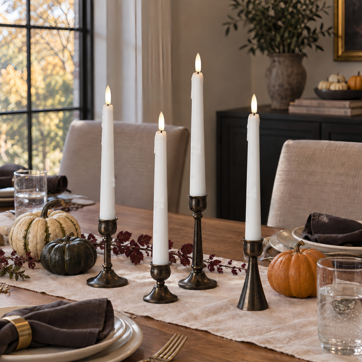 Four white taper candles in dark metal candlesticks arranged on a dining table runner, with pumpkins, plates, and a home dining room background.