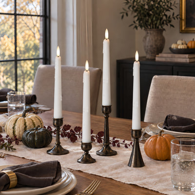 Four white taper candles in dark metal candlesticks arranged on a dining table runner, with pumpkins, plates, and a home dining room background.